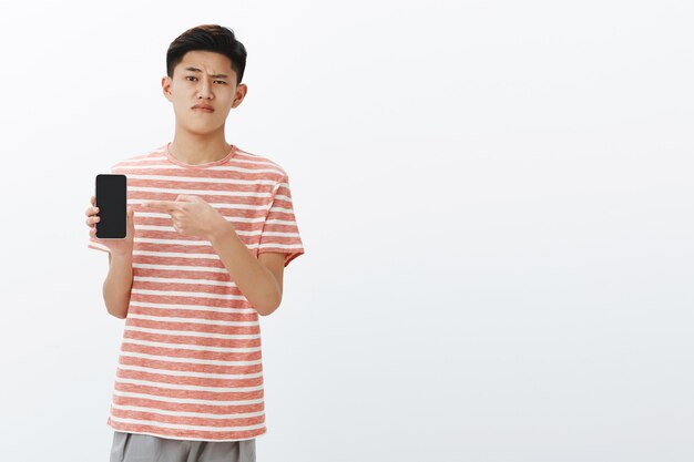 Unimpressed displeased and indifferent asian smart male student holding smartphone and pointing at device screen raising one eyebrow in doubt, pursing lips