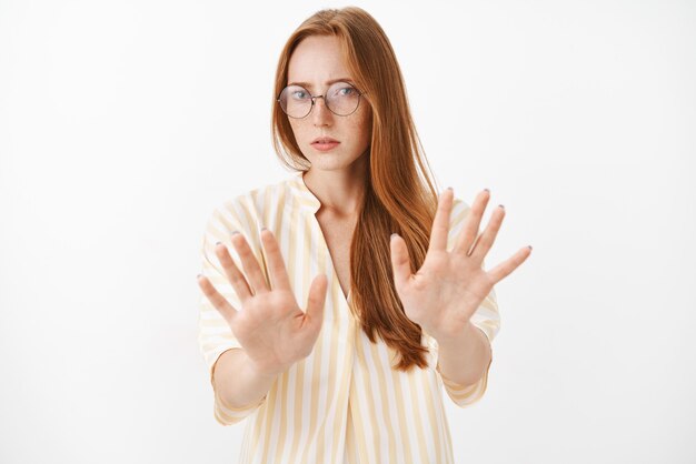 Unimpressed and dissatisfied bossy female journalist in trendy glasses with freckles pulling palms towards in rejection or stop gesture disliking offer gazing with aversion