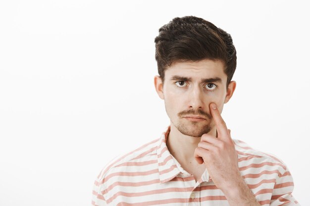 Unimpressed with boring conversation. Displeased bored guy in casual striped shirt, pulling eye and showing eyelid with indifferent careless expression, standing gloomy and calm over gray wall