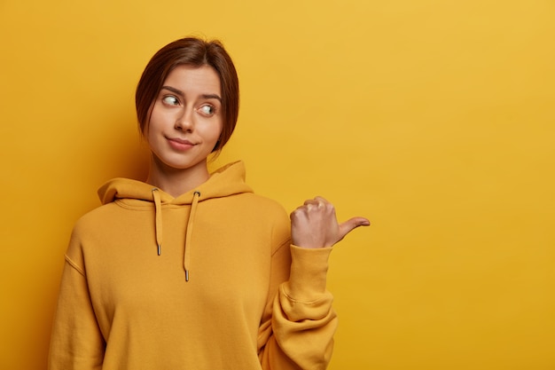 Unimpressed woman points thumb aside, explains something and gives direction, has combed hair, wears yellow hoodie, says check out, has serious displeased expression