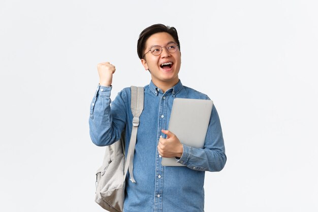 Университет, учеба за границей и концепция образа жизни. Счастливый радующийся азиатский студент-мужчина с триумфом подтяжек, сдающий экзамены, заканчивающий последний семестр, кулаками и удовлетворенный криком «да», держите ноутбук.