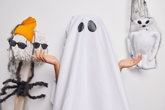 Unknown female ghost coned with white sheet spreads palms with hesitation tries to look spooky wears ghost costume celebrates halloween poses indoor. Party celebration and mystery concept.