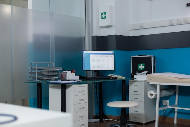 Unoccupied modern clinic workspace with nobody in it having turned on computer screen showing medical report. Medical room having firstaid kit, contemporary hospital furniture and patients documents.