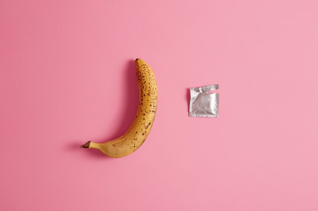Unpacked condom and one whole banana on pink studio background. 