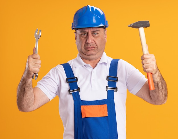 Unpleased adult builder man in uniform holds wrench and hammer isolated on orange wall