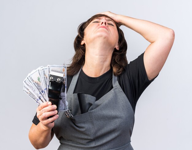 Unpleased adult caucasian female barber in uniform putting hand on forehead and holding money with hair clipper isolated on white background with copy space