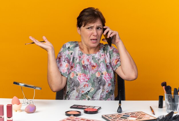 電話で話している化粧ツールと横を見て化粧ブラシを持ってテーブルに座っている不機嫌な大人の白人女性