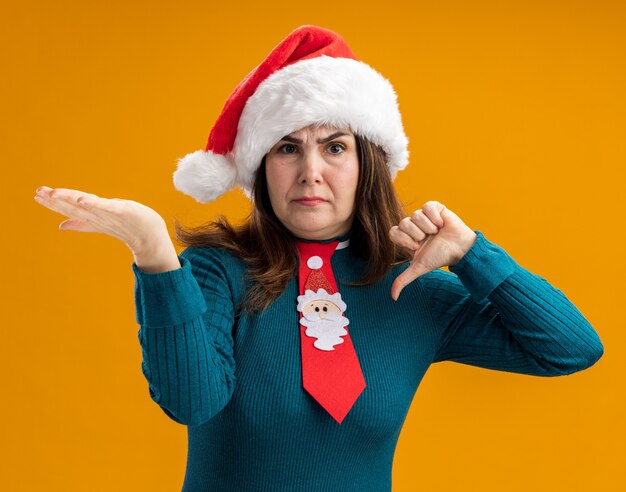 Unpleased adult caucasian woman with santa hat and santa tie thumbs down and keeps hand open isolated on orange background with copy space