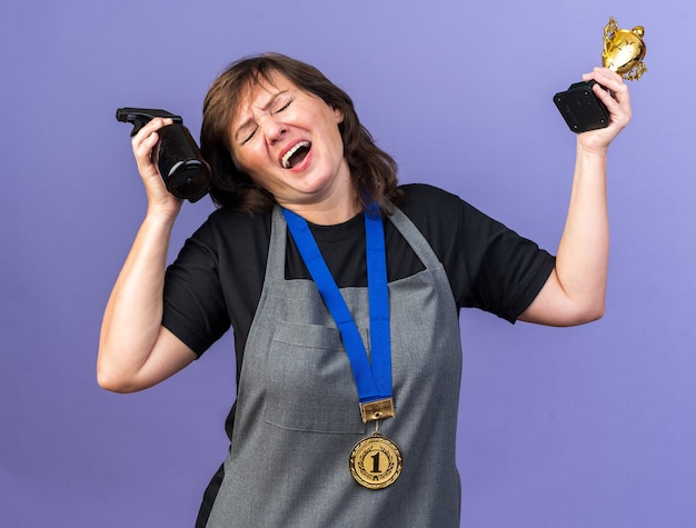 unpleased adult female barber in uniform with golden medal around neck holding spray bottle and winner cup isolated on purple wall with copy space