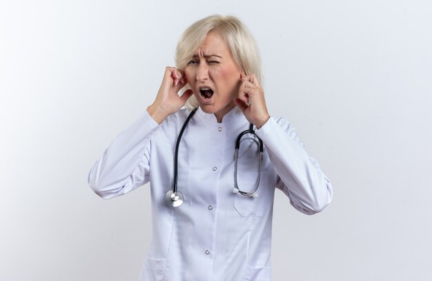 의료 가운을 입은 불쾌한 성인 여성 의사, 복사 공간이 있는 흰 벽에 격리된 손가락으로 귀를 닫는 청진기