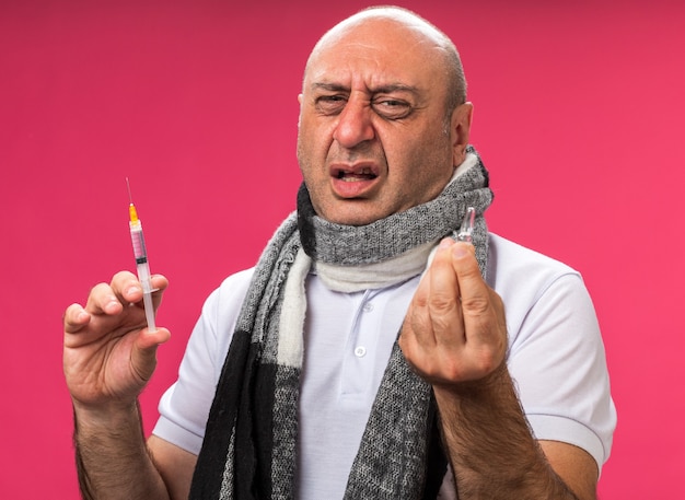 unpleased adult ill caucasian man with scarf around neck holding syringe and ampoule isolated on pink wall with copy space