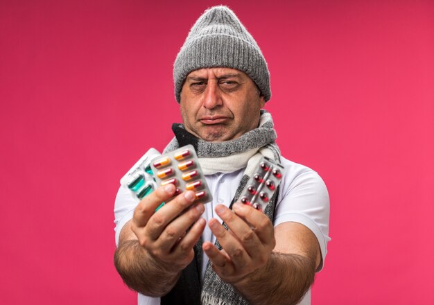 unpleased adult ill caucasian man with scarf around neck wearing winter hat holding different medicine packs isolated on pink wall with copy space