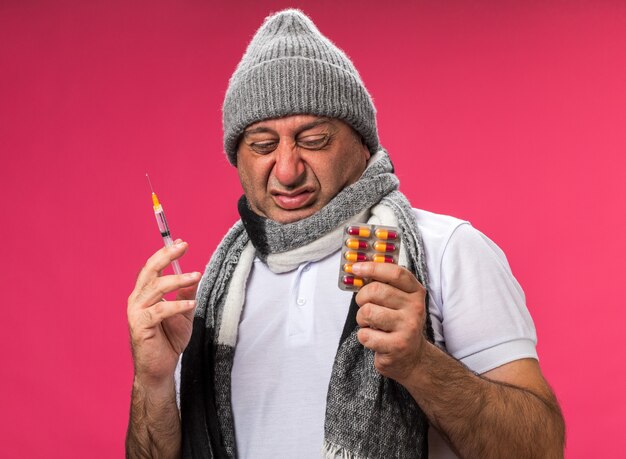 unpleased adult ill caucasian man with scarf around neck wearing winter hat holding syringe and looking at medicine blister pack isolated on pink wall with copy space