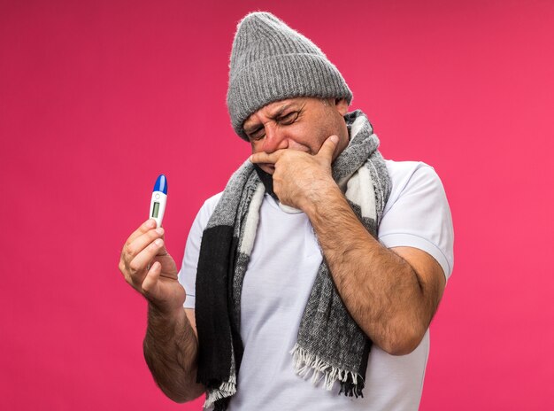 unpleased adult ill caucasian man with scarf around neck wearing winter hat puts hand on chin holding and looking at thermometer isolated on pink wall with copy space