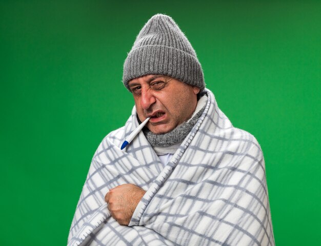 首の周りにスカーフを身に着けている不機嫌な大人の病気の白人男性は、コピースペースのある緑の壁に隔離された彼の口の中に温度計を保持している格子縞に包まれた冬の帽子をかぶっています