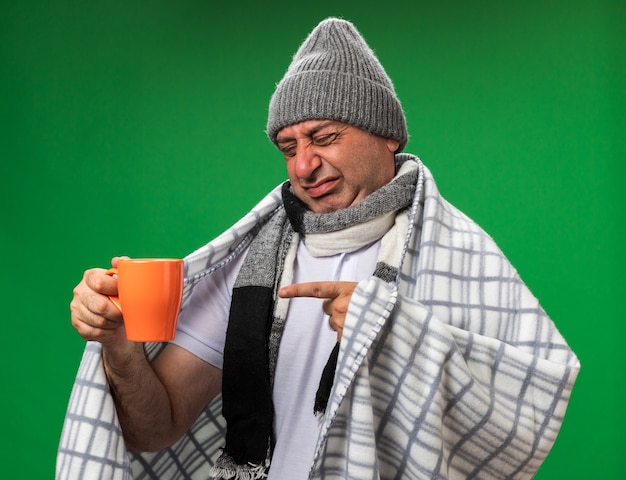 unpleased adult ill caucasian man with scarf around neck wearing winter hat wrapped in plaid looking and pointing at cup isolated on green wall with copy space