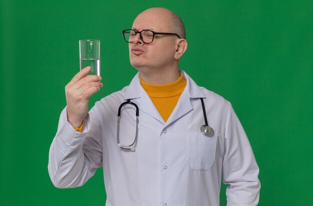 Недовольный взрослый мужчина в очках в униформе врача со стетоскопом, держащим и смотрящим на стакан воды