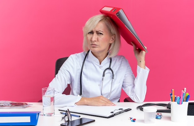 Unpleased adult slavic female doctor in medical robe with stethoscope sitting at desk with office tools holding file folder isolated on pink background with copy space