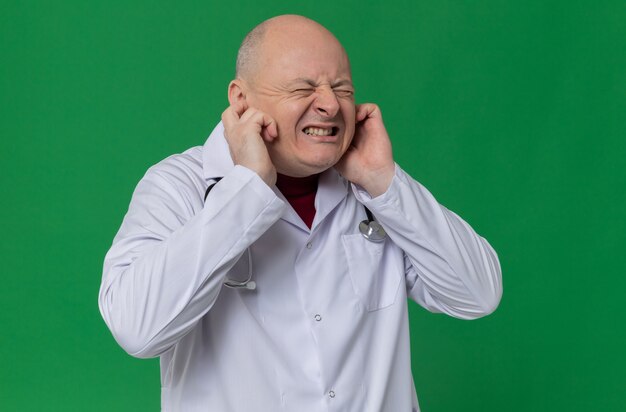 Unpleased adult slavic man in doctor uniform with stethoscope closing his ears with fingers