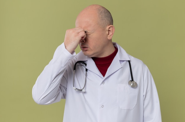 Uomo slavo adulto scontento in uniforme da medico con uno stetoscopio che si tiene il naso