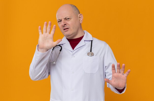 Unpleased adult slavic man in doctor uniform with stethoscope keeping hands open gesturing stop sign