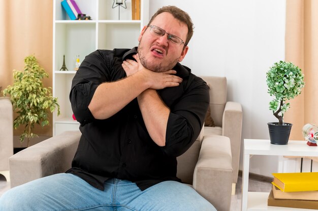 Unpleased adult slavic man in optical glasses sits on armchair pretending to choke himself inside living room