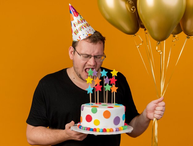 Unpleased adult slavic man in optical glasses wearing birthday cap holds helium balloons and birthday cake 