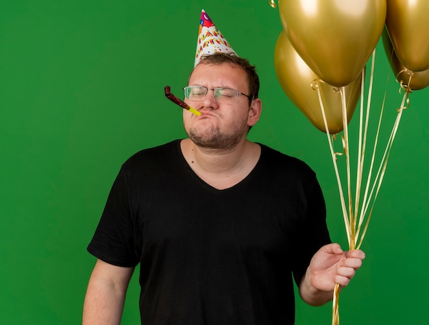 Недовольный взрослый славянский мужчина в оптических очках и кепке на день рождения держит гелиевые шары, дует в свисток