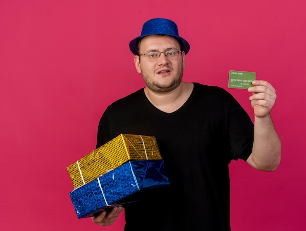 Unpleased adult slavic man in optical glasses wearing blue party hat holds gift boxes and credit card 