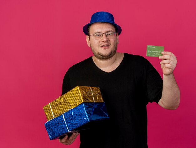 青いパーティー ハットを身に着けている光学メガネで不愉快な大人のスラブ人は、ギフト ボックスとクレジット カードを保持します。