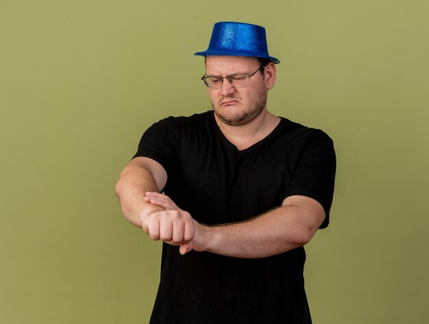 Unpleased adult slavic man in optical glasses wearing blue party hat holds and looks at arm 