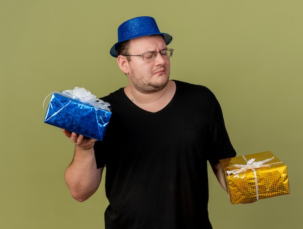 Недовольный взрослый славянский мужчина в оптических очках в синей праздничной шляпе держит и смотрит на подарочные коробки