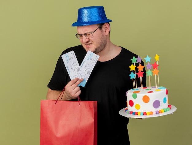 L'uomo slavo adulto scontento con gli occhiali ottici che indossa un cappello da festa blu tiene la torta di compleanno della borsa della spesa di carta e i biglietti aerei