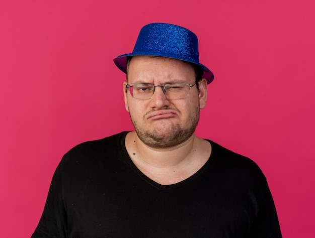 Unpleased adult slavic man in optical glasses wearing blue party hat looks at camera 