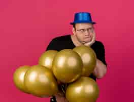 Free photo unpleased adult slavic man in optical glasses wearing blue party hat pus hand on chin and holds helium balloons looking at side