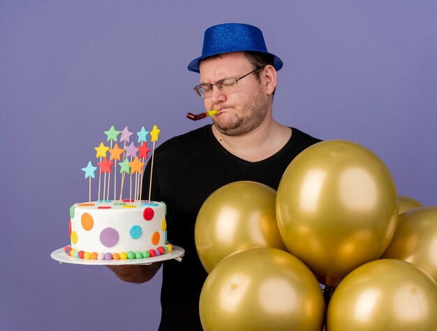 Unpleased adult slavic man in optical glasses wearing blue party hat stands with helium balloons holding birthday cake and blowing party whistle 