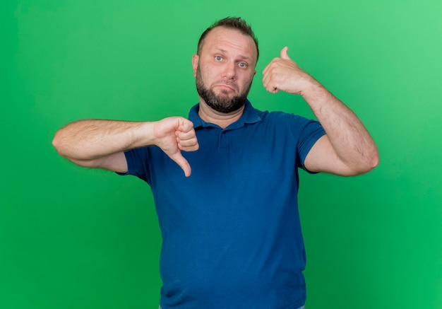 Free photo unpleased adult slavic man showing thumbs up and down  isolated on green wall