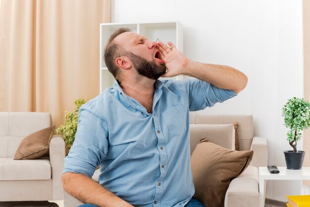 Unpleased adult slavic man sits on armchair holding hand close to mouth looking at side pretending to call someone inside the living room