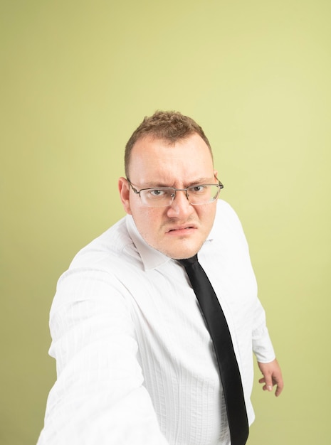 Бесплатное фото Недовольный взрослый славянский мужчина в очках и галстуке смотрит в камеру, протягивая руку к камере, изолированной на оливково-зеленом фоне