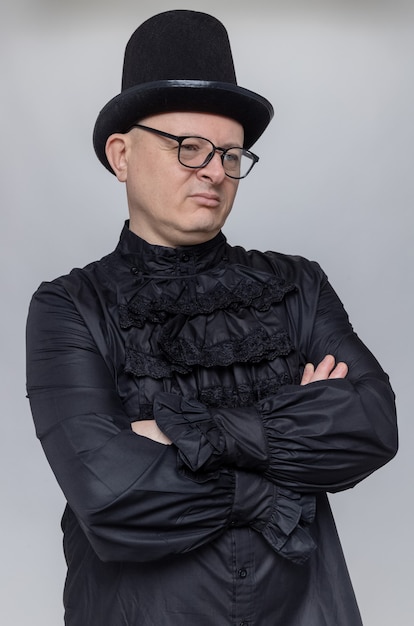 Unpleased adult slavic man with top hat and optical glasses in black gothic shirt standing with crossed arms and looking at side 