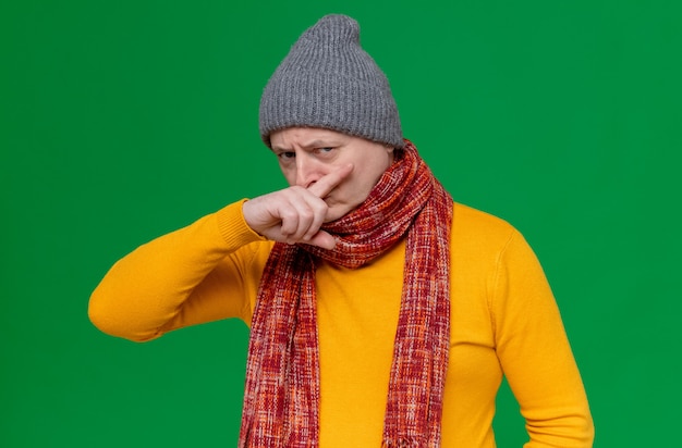 Unpleased adult slavic man with winter hat and scarf around his neck wipes his nose with finger looking at front