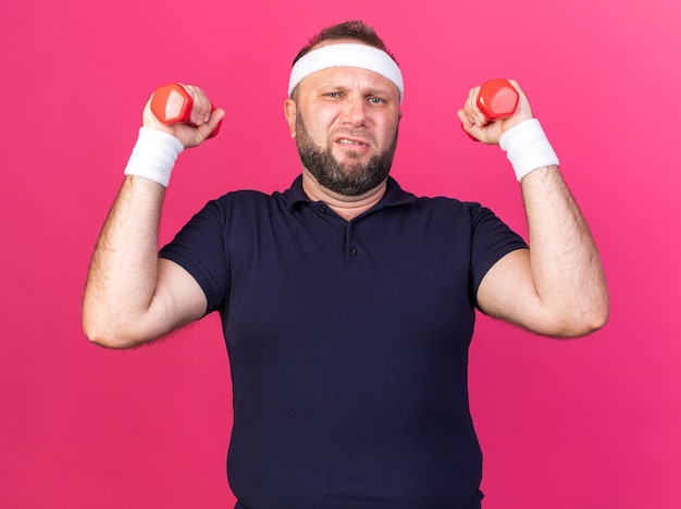 unpleased adult slavic sporty man wearing headband and wristbands holding dumbbells isolated on pink wall with copy space