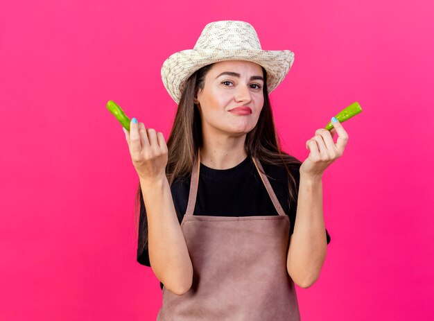 Unpleased beautiful gardener girl in uniform wearing gardening hat holding a broken pepper isolated on pink background