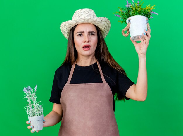 Unpleased beautiful gardener girl in uniform wearing gardening hat holding flowers in flowerpot isolated on green background