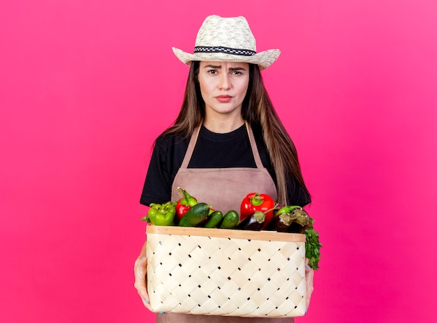ピンクの背景に分離された野菜のバスケットを保持しているガーデニング帽子を身に着けている制服を着た不機嫌な美しい庭師の女の子