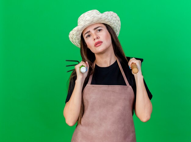 Unpleased beautiful gardener girl in uniform wearing gardening hat putting rake with hoe rake on shoulder isolated on green