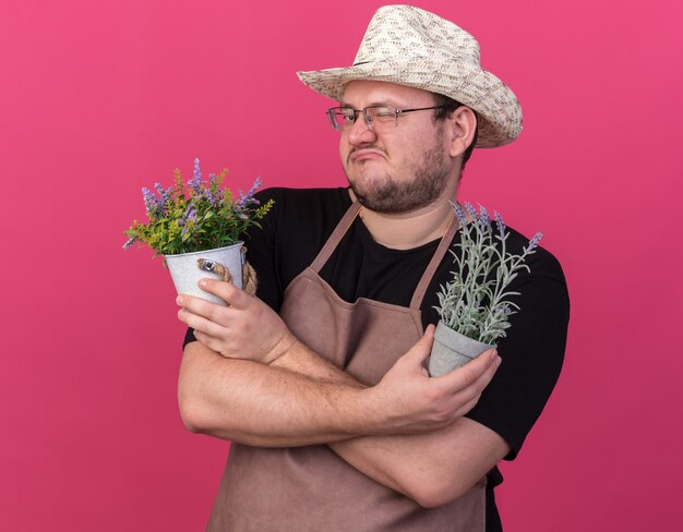 Unpleased blinked young male gardener wearing gardening hat holding and crossing flowers in flowerpots isolated on pink wall