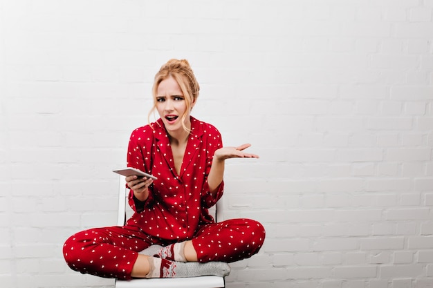 スマートフォンを持っている不機嫌なブロンドの女の子。明るい壁に足を組んで座っている赤いパジャマの怒っている女性モデル。