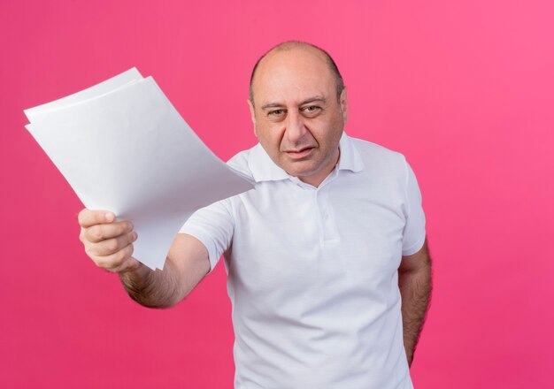 Unpleased casual mature businessman stretching out documents at camera isolated on pink background