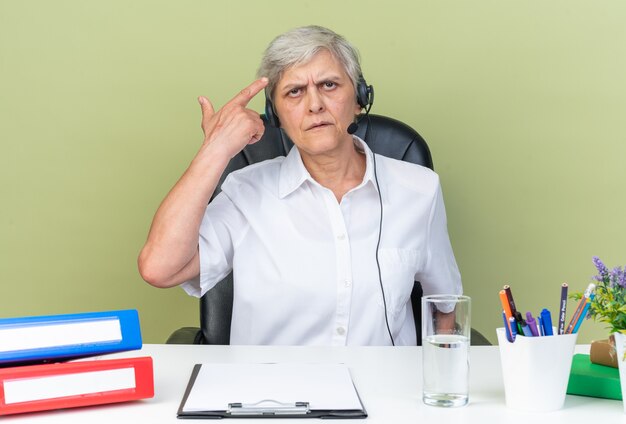 Unpleased caucasian female call center operator on headphones sitting at desk with office tools pointing at her hair